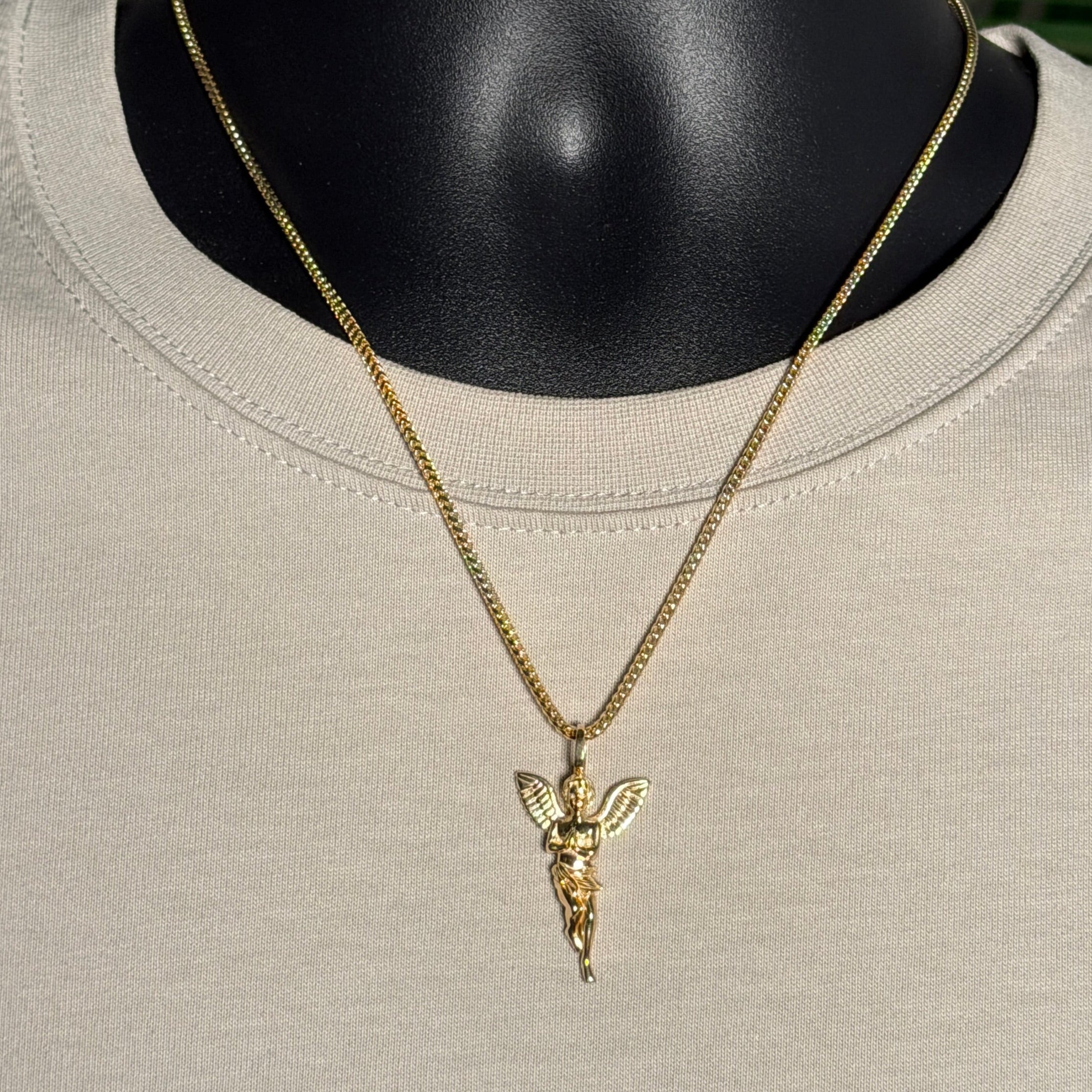 A gold chain necklace with a winged cherub pendant displayed on a black mannequin torso wearing a light beige crewneck shirt, with blurred green leaves in the background.