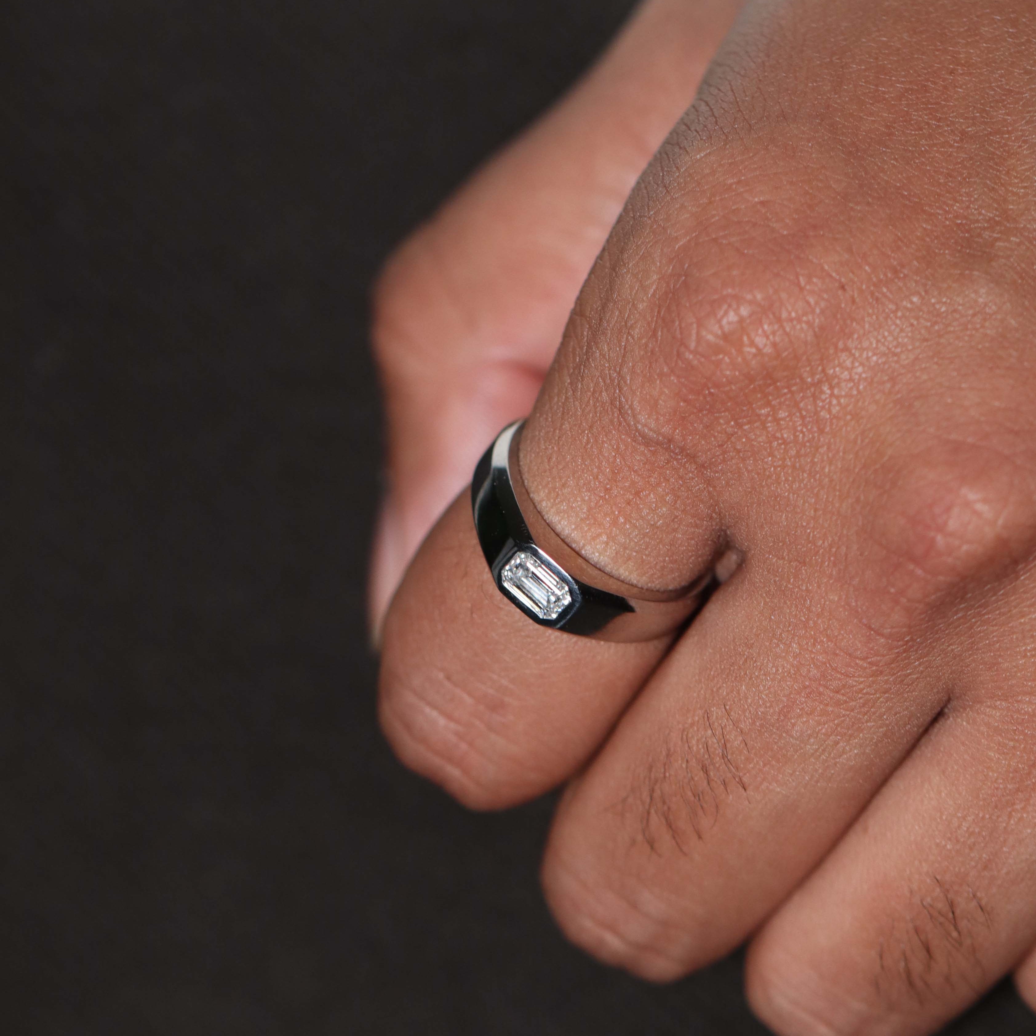 A close-up of a mans hand wearing a platinum-toned metal ring with a rectangular emerald diamond on a dark background.