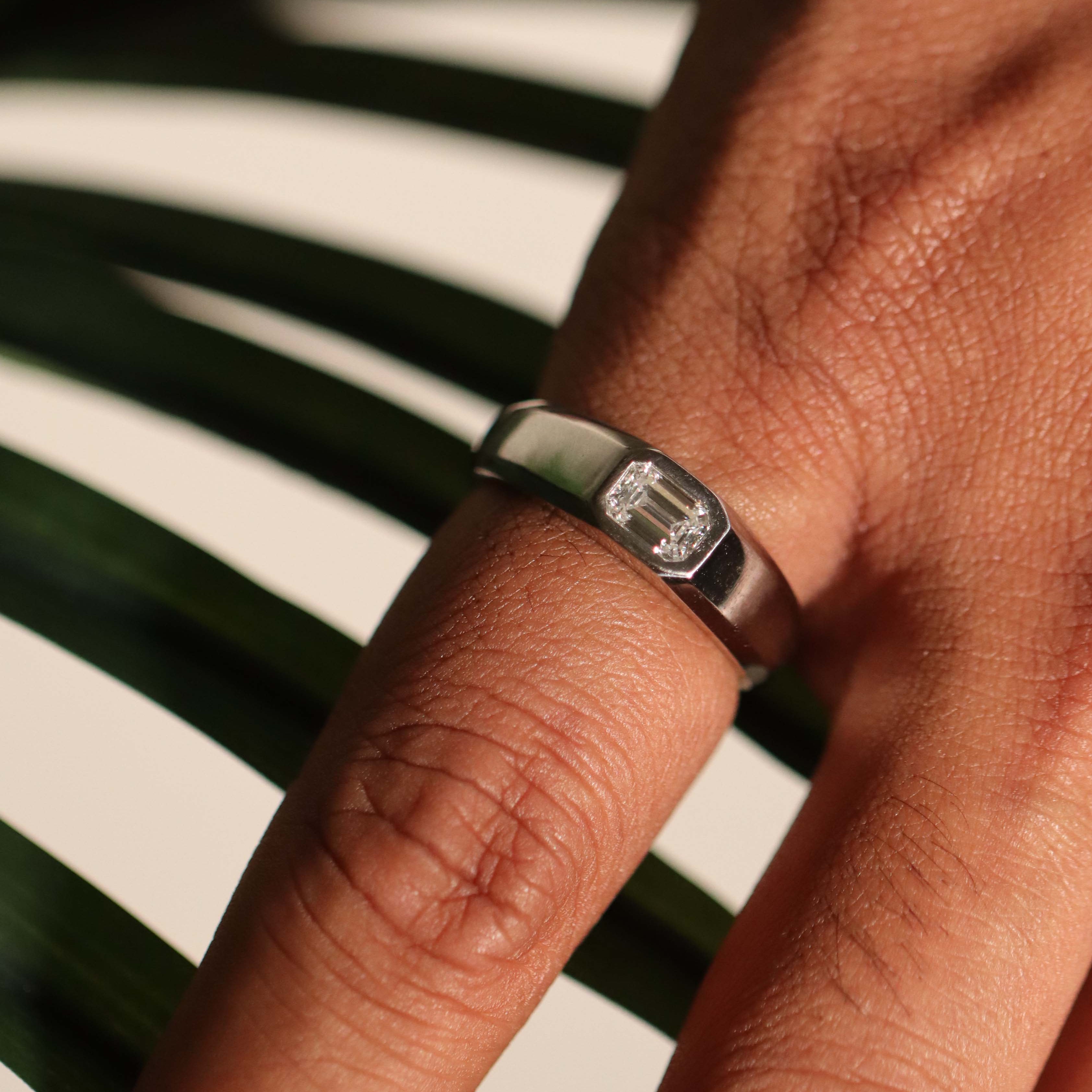 A close-up of a silver ring with an emerald-cut clear gemstone worn on a person's finger, set against a background of green palm leaves and a light beige surface.